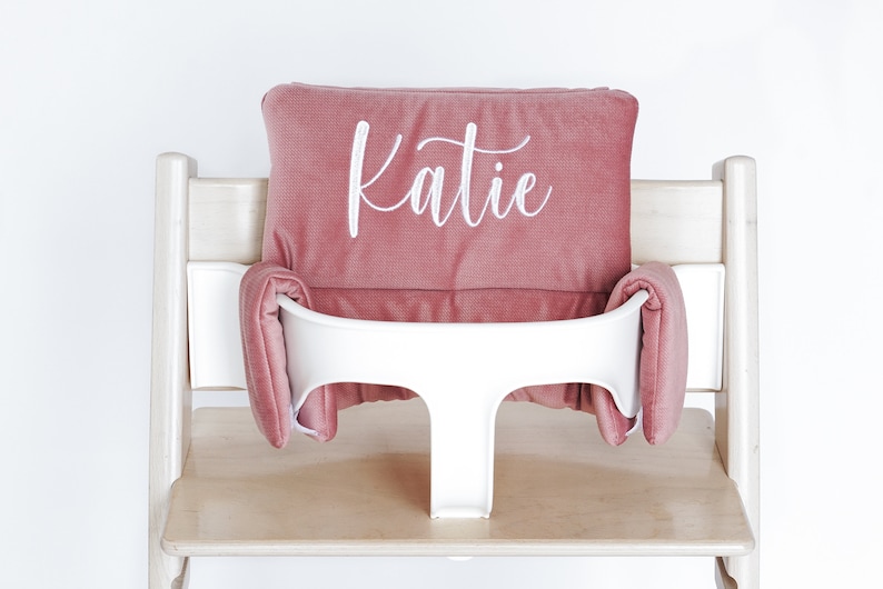 May include: A dusty rose high chair cushion set with the name "Katie" embroidered in white script. The set includes a headrest and side cushions, designed to fit a high chair. The high chair is made of light wood and has a white plastic seat.