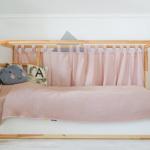 May include: A wooden bed frame with a pink checkered blanket and pink curtains. The bed has a white base and a gray cloud-shaped pillow with a smiling face. There is a white rabbit figurine on the left side of the bed.