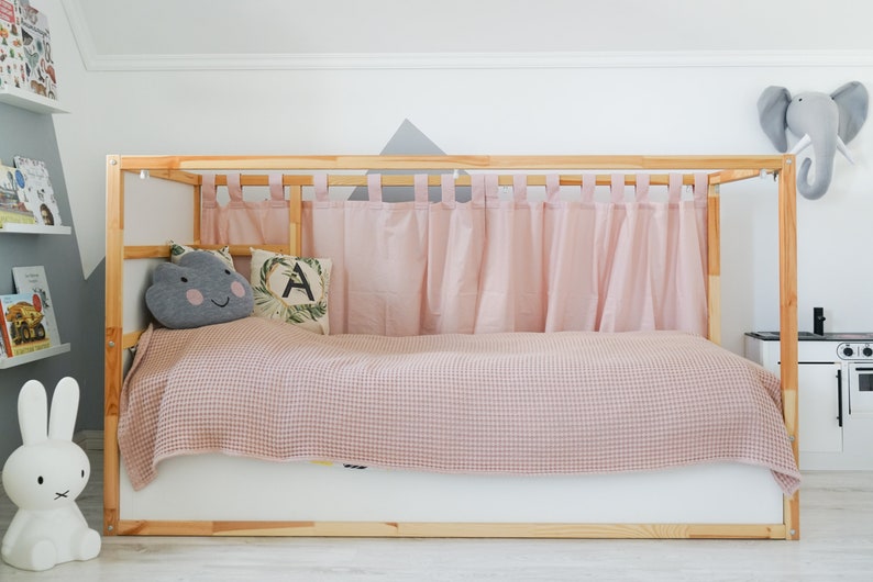 May include: A wooden bed frame with a pink checkered blanket and pink curtains. The bed has a white base and a gray cloud-shaped pillow with a smiling face. There is a white rabbit figurine on the left side of the bed.