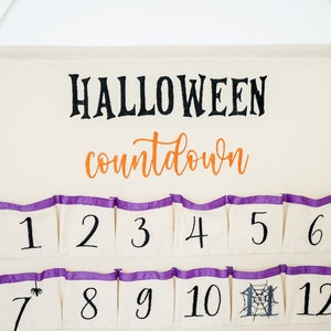 May include: A cream-colored Halloween countdown calendar with pockets numbered 1-12. The word "HALLOWEEN" is in black script above "countdown" in orange script. Each pocket has a purple trim.