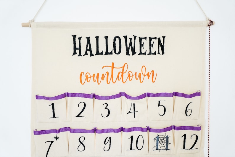 May include: A cream-colored Halloween countdown calendar with pockets numbered 1-12. The word "HALLOWEEN" is in black script above "countdown" in orange script. Each pocket has a purple trim.