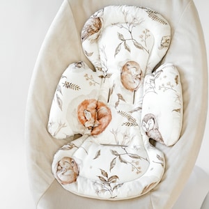 Op de afbeelding: Een witte en beige baby schommelstoel met een uitneembare, gevoerde inzet met een bosdierprint. De inzet heeft een witte achtergrond met bruine en oranje vossen, uilen en bladeren.