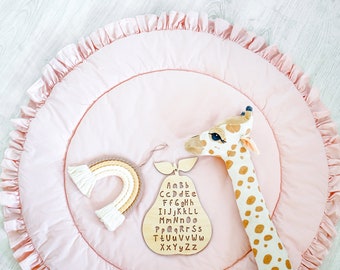 May include: A pink play mat with a ruffled edge, featuring a wooden pear with the alphabet, a rainbow-shaped toy, and a stuffed giraffe.