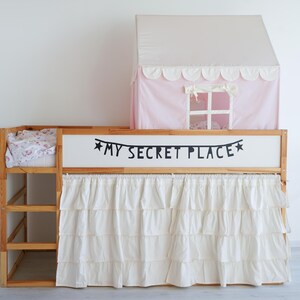Pode incluir: Uma cama de beliche de madeira com uma cortina branca com babados e uma estrutura de tenda rosa no topo. A cama tem uma cabeceira branca com letras pretas que dizem "MY SECRET PLACE" com estrelas de cada lado.