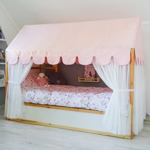 May include: A pink canopy bed with white curtains and a floral patterned bedspread. The bed is made of wood and has a white base. There are bookshelves on the wall behind the bed.