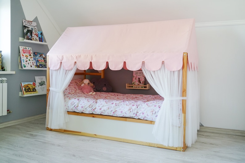 May include: A pink canopy bed with white curtains and a floral patterned bedspread. The bed is made of wood and has a white base. There are bookshelves on the wall behind the bed.