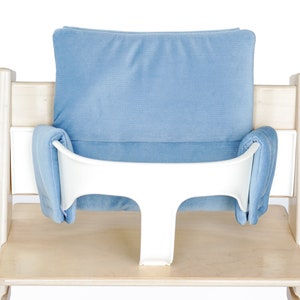 May include: A light wood high chair with a blue cushion and a white safety bar. The chair has a light blue padded backrest and seat inserts. The safety bar is white and T-shaped.