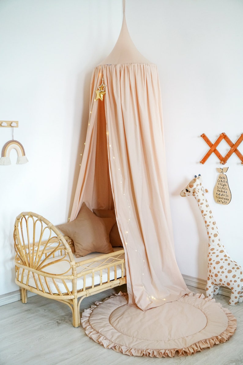 May include: A light pink canopy bed with a white wicker bed frame and a light pink ruffled rug. The canopy is decorated with a string of fairy lights and a gold star.