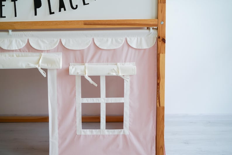 May include: A pink fabric canopy with white scalloped trim and two white fabric window coverings. The canopy is attached to a wooden bed frame.