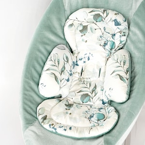 Op de afbeelding: Een lichtgroene baby schommel met een wit en groen bloemmotief kussen. Het kussen heeft een verhoogde hoofdsteun en een gecontourde vorm voor comfort.