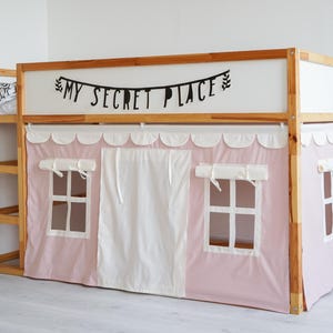 May include: A wooden bunk bed with a pink and white tent-like canopy. The canopy has two windows and a white curtain in the middle. The bed has a wooden ladder and a shelf on the side. The top of the bed has a sign that says "My Secret Place".