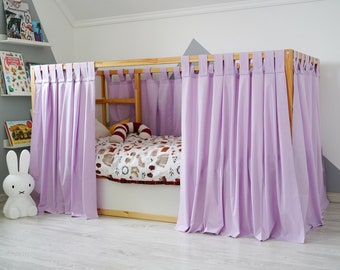 May include: A wooden bed frame with lavender curtains draped around it. The bed is made with a white and brown patterned bedding and a red and white striped pillow. 