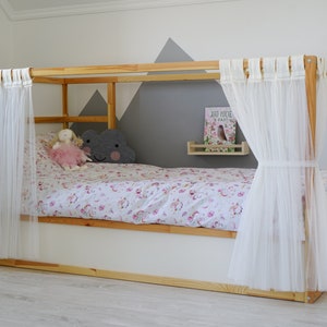 May include: A wooden bed frame with white sheer curtains, a white mattress, and a pink and white floral duvet cover. A gray cloud-shaped pillow and a pink doll are on the bed. A small shelf with a book titled "Jod Mocne i Szybkie" is on the wall behind the bed.