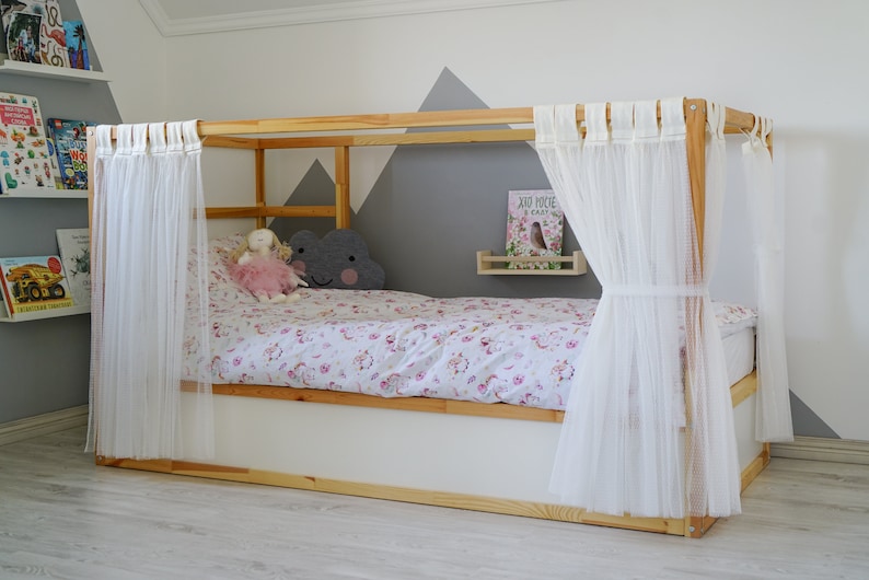 May include: A wooden bed frame with white sheer curtains, a white mattress, and a pink and white floral duvet cover. A gray cloud-shaped pillow and a pink doll are on the bed. A small shelf with a book titled "Jod Mocne i Szybkie" is on the wall behind the bed.