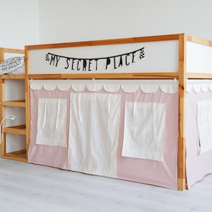 May include: A wooden loft bed with a pink fabric canopy and white curtains. The bed has a shelf on the side and a black banner that reads "My Secret Place".