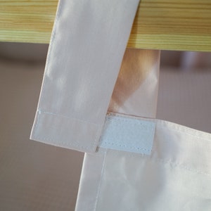 May include: A close-up of a pink fabric curtain with a white velcro strip attached to the top edge. The curtain is hanging from a wooden rod.