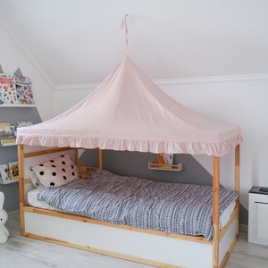 May include: A light pink fabric canopy with a ruffled edge suspended over a wooden bed frame. The bed features a white base, a mattress with a black and white patterned duvet cover, and a white pillow with black polka dots.