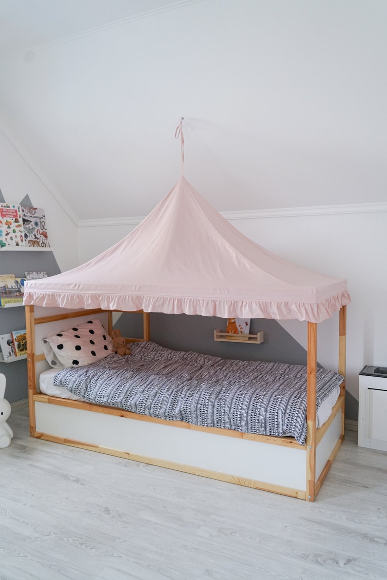 May include: A light pink fabric canopy with a ruffled edge suspended over a wooden bed frame. The bed features a white base, a mattress with a black and white patterned duvet cover, and a white pillow with black polka dots.