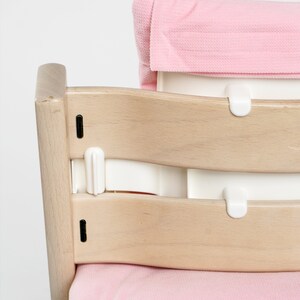 Op de afbeelding: Een houten hoge stoel met een roze gepolsterde zitting en rugleuning. De stoel heeft een witte plastic band die aan de achterkant van de stoel wordt bevestigd.