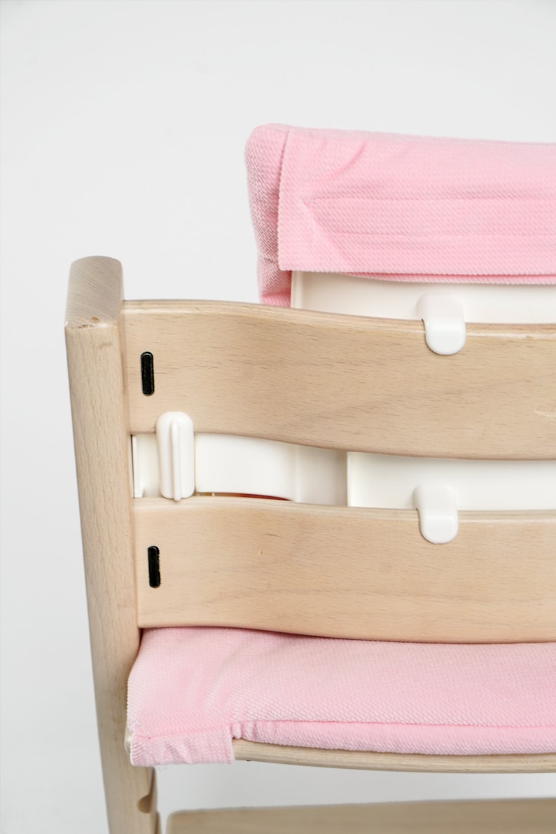 Op de afbeelding: Een houten hoge stoel met een roze gepolsterde zitting en rugleuning. De stoel heeft een witte plastic band die aan de achterkant van de stoel wordt bevestigd.