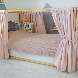 May include: A wooden bed frame with pink curtains draped around it. The bed has a pink and white checkered blanket and two pillows. One pillow has a gray cloud with a smiley face on it and the other pillow has a green leaf with the letter A on it.