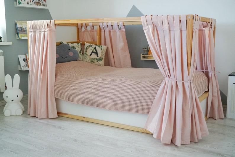 May include: A wooden bed frame with pink curtains draped around it. The bed has a pink and white checkered blanket and two pillows. One pillow has a gray cloud with a smiley face on it and the other pillow has a green leaf with the letter A on it.