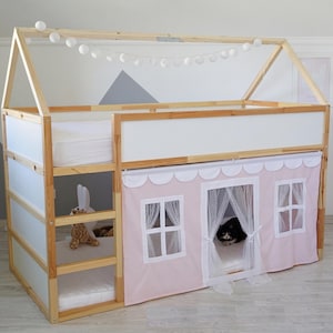 May include: A wooden house-shaped loft bed with a pink and white fabric house facade. The bed has a ladder, a lower bunk, and a top bunk. A string of white globe lights hangs above the bed. A cat is visible inside the house.