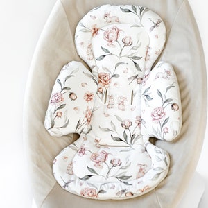 Op de afbeelding: Een baby wipstoel zitkussen met een bloemenpatroon van roze rozen, groene bladeren en kleine konijntjes op een witte achtergrond. Het kussen is geplaatst tegen een neutraal gekleurd wipstoel frame.