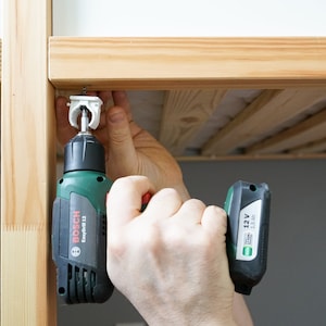 Puede incluir: Una persona utiliza un taladro Bosch EasyDrill 12 verde y negro para sujetar un soporte met&aacute;lico blanco a un marco de cama de madera.