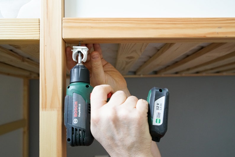 Puede incluir: Una persona utiliza un taladro Bosch EasyDrill 12 verde y negro para sujetar un soporte met&aacute;lico blanco a un marco de cama de madera.
