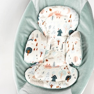 Op de afbeelding: Een groene baby schommel met een witte en bruine gepatroneerde inzet. De inzet heeft een bos thema met bomen, paddenstoelen en dieren.