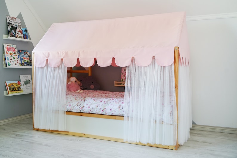 May include: A pink and white canopy bed with a scalloped edge and sheer curtains. The bed is made with a floral patterned bedding and has a wooden frame.