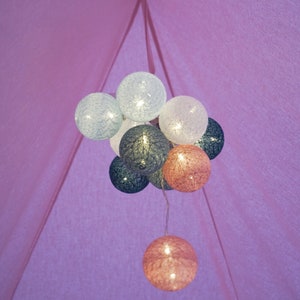 May include: A cluster of spherical string lights hanging inside a pink fabric tent. The illuminated globes feature a textured, woven thread design in shades of white, light blue, dark blue, and soft pink, creating a glowing decorative effect.