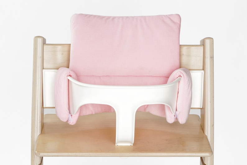 Op de afbeelding: Een roze gevoerd hoge stoel kussen met een witte lade. Het kussen is gemaakt van zachte stof en heeft een comfortabel ontwerp.