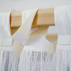 May include: White curtains with a lace trim hang from a wooden rod. The curtains have a white fabric top with a hook and loop fastener.
