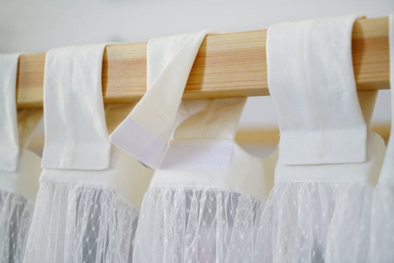 May include: White curtains with a lace trim hang from a wooden rod. The curtains have a white fabric top with a hook and loop fastener.