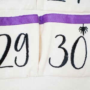 May include: Close-up of a cream-colored fabric advent calendar with black embroidered numbers 29 and 30. The pockets have a purple ribbon trim. A black spider is embroidered above the number 30.
