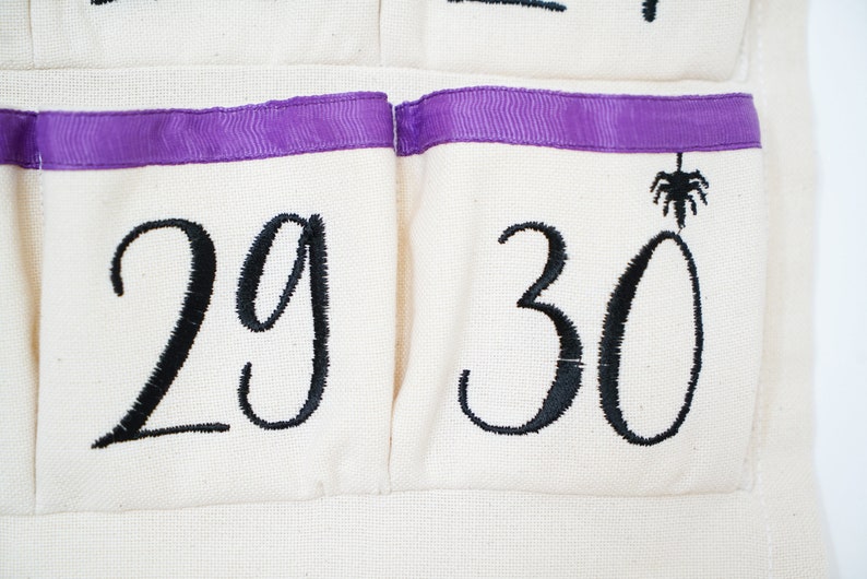 May include: Close-up of a cream-colored fabric advent calendar with black embroidered numbers 29 and 30. The pockets have a purple ribbon trim. A black spider is embroidered above the number 30.