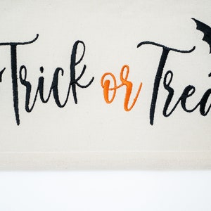 May include: A cream-colored Halloween banner with the words "Trick or Treat" embroidered in black and orange script. Black bat silhouettes are embroidered on either side of the text, creating a festive and spooky design.