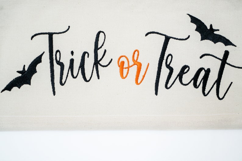 May include: A cream-colored Halloween banner with the words "Trick or Treat" embroidered in black and orange script. Black bat silhouettes are embroidered on either side of the text, creating a festive and spooky design.