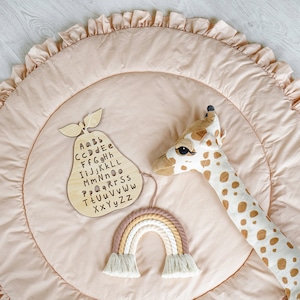 May include: A light pink play mat with a ruffled edge. The mat features a wooden pear-shaped alphabet chart, a rainbow-colored macrame wall hanging, and a stuffed giraffe toy.