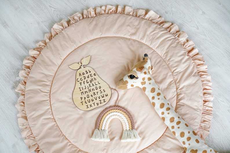 May include: A light pink play mat with a ruffled edge. The mat features a wooden pear-shaped alphabet chart, a rainbow-colored macrame wall hanging, and a stuffed giraffe toy.