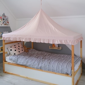 May include: A light pink fabric bed canopy with a ruffled edge, suspended from the ceiling over a wooden frame bed. The bed features a white and black patterned duvet cover, a white pillow with black polka dots, and a small brown plush toy.