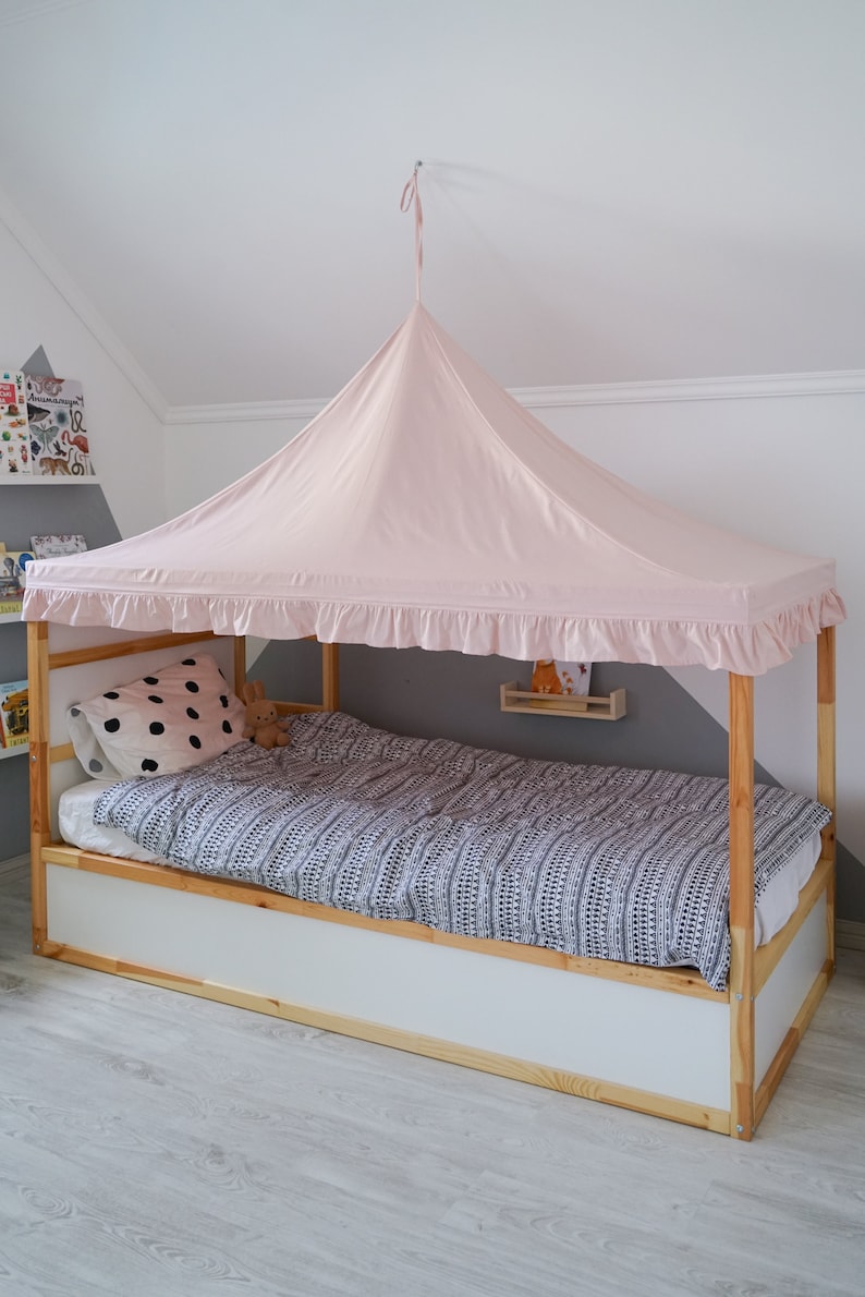 May include: A light pink fabric bed canopy with a ruffled edge, suspended from the ceiling over a wooden frame bed. The bed features a white and black patterned duvet cover, a white pillow with black polka dots, and a small brown plush toy.