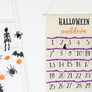 May include: A Halloween countdown calendar with 30 numbered pockets, purple trim, and a hanging bat and spider decoration. A garland with orange circles, bats, and spiders hangs on a white ladder.