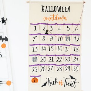 May include: A Halloween countdown calendar made of beige fabric with purple trim and pockets. The calendar has the words "HALLOWEEN countdown" in black and orange. Numbers 1-31 are printed on the pockets, with Halloween-themed illustrations. The words "Trick or Treat" are also printed on the calendar.