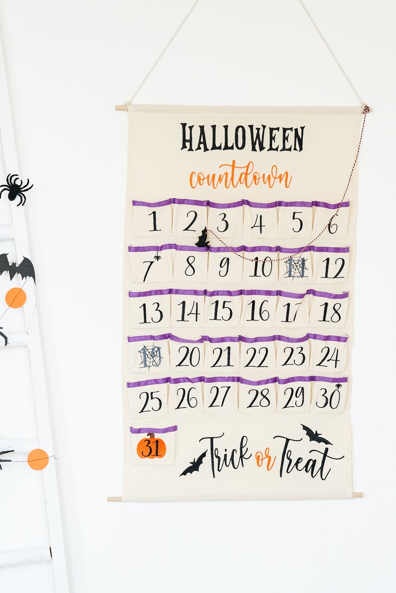 May include: A Halloween countdown calendar made of beige fabric with purple trim and pockets. The calendar has the words "HALLOWEEN countdown" in black and orange. Numbers 1-31 are printed on the pockets, with Halloween-themed illustrations. The words "Trick or Treat" are also printed on the calendar.
