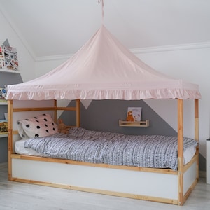 Puede incluir: Una cama de madera blanca con una tienda de campaña de dosel rosa drapeada sobre ella. La cama tiene una almohada blanca con lunares negros y una manta de patrón gris y blanco. Hay un pequeño estante en la pared sobre la cama con un libro encima.