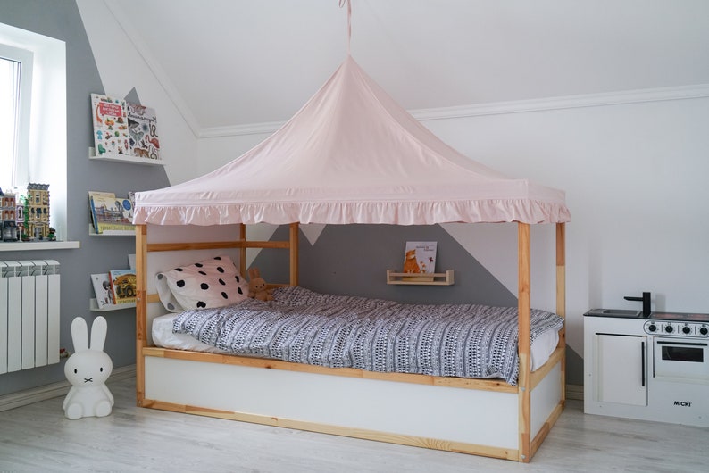 May include: A wooden bed frame with a white base and a light pink fabric canopy featuring a ruffled edge. The bed is dressed with a gray and white patterned duvet cover and a white pillow with black polka dots, creating a cozy sleeping space.