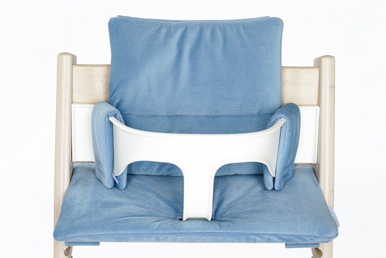 May include: A light wood high chair with a blue cushion and backrest. The chair has a white safety bar. The cushion and backrest are a solid light blue color. The chair is designed for a baby or toddler.